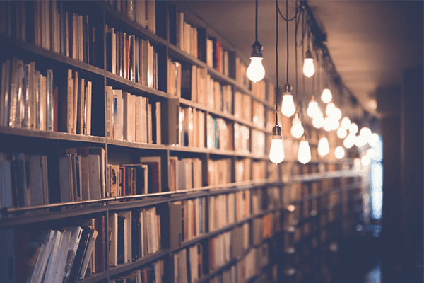Rows of books fill tall shelves in a library with warm, exposed light bulbs hanging from the ceiling, creating a cozy and inviting atmosphere.