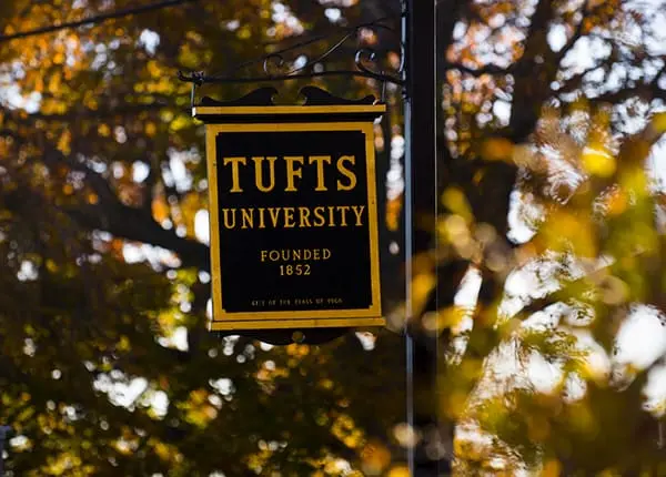 A black and gold sign reads Tufts University Founded 1852 and hangs outdoors with autumn-colored trees blurred in the background.