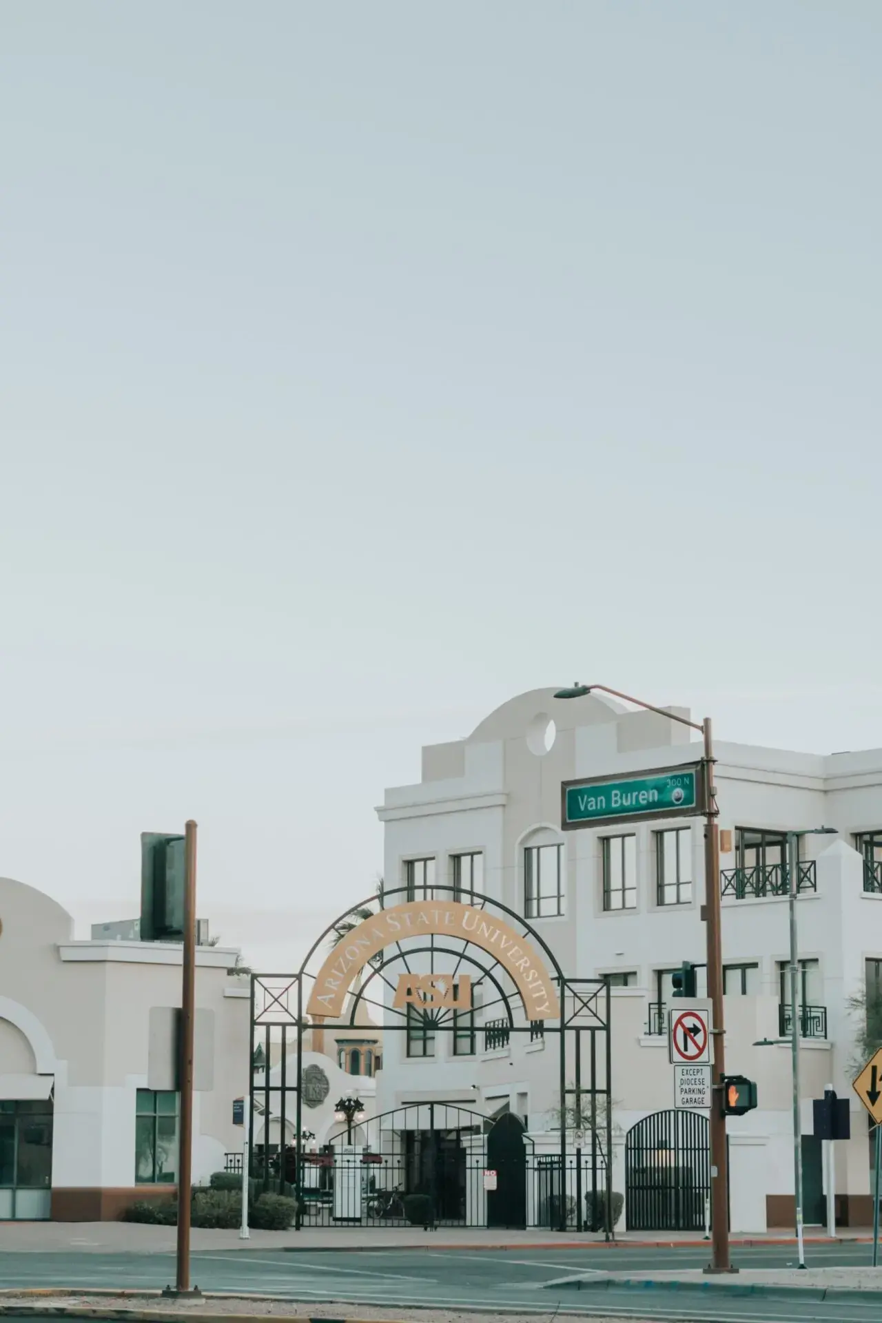 A large arched gate reading Arizona State University stands in front of a white building with art deco architecture. A street sign for Van Buren St is visible, along with traffic signals and a clear sky overhead.