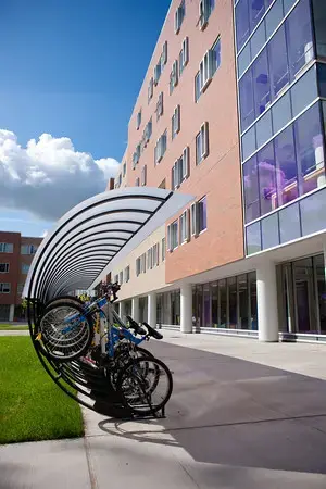 Several bicycles are parked in a covered bike rack outside a modern multi-story building with large windows on a sunny day. The sky is blue with some fluffy clouds.