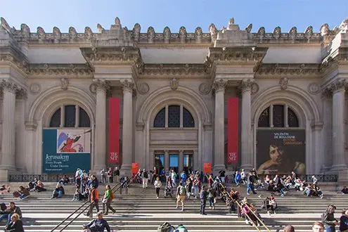 People sit and walk on the wide steps leading to the entrance of the Metropolitan Museum of Art in New York City, with large columns, arched windows, and banners promoting exhibitions.
