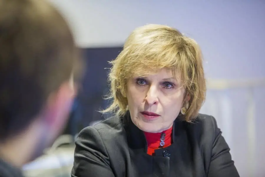 A woman with short blonde hair and a serious expression wears a black jacket over a red top, sitting indoors and speaking to someone whose back is to the camera.