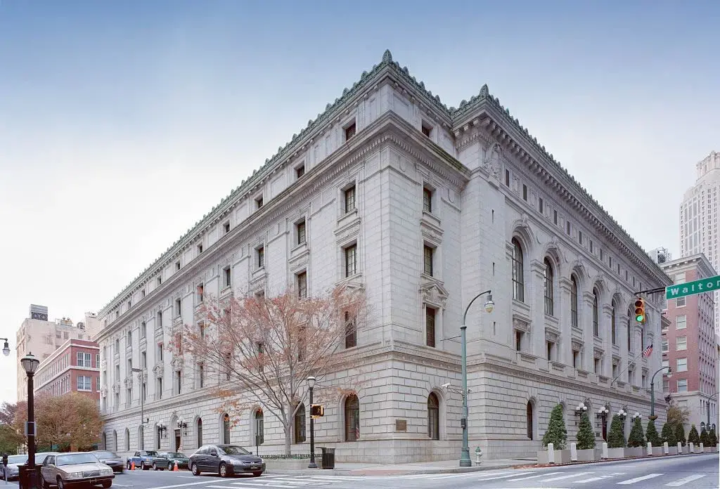 A large, historic, white stone building with ornate architectural details and arched windows sits on a street corner under a clear sky. Leafless trees and traffic lights are visible along the sidewalk.