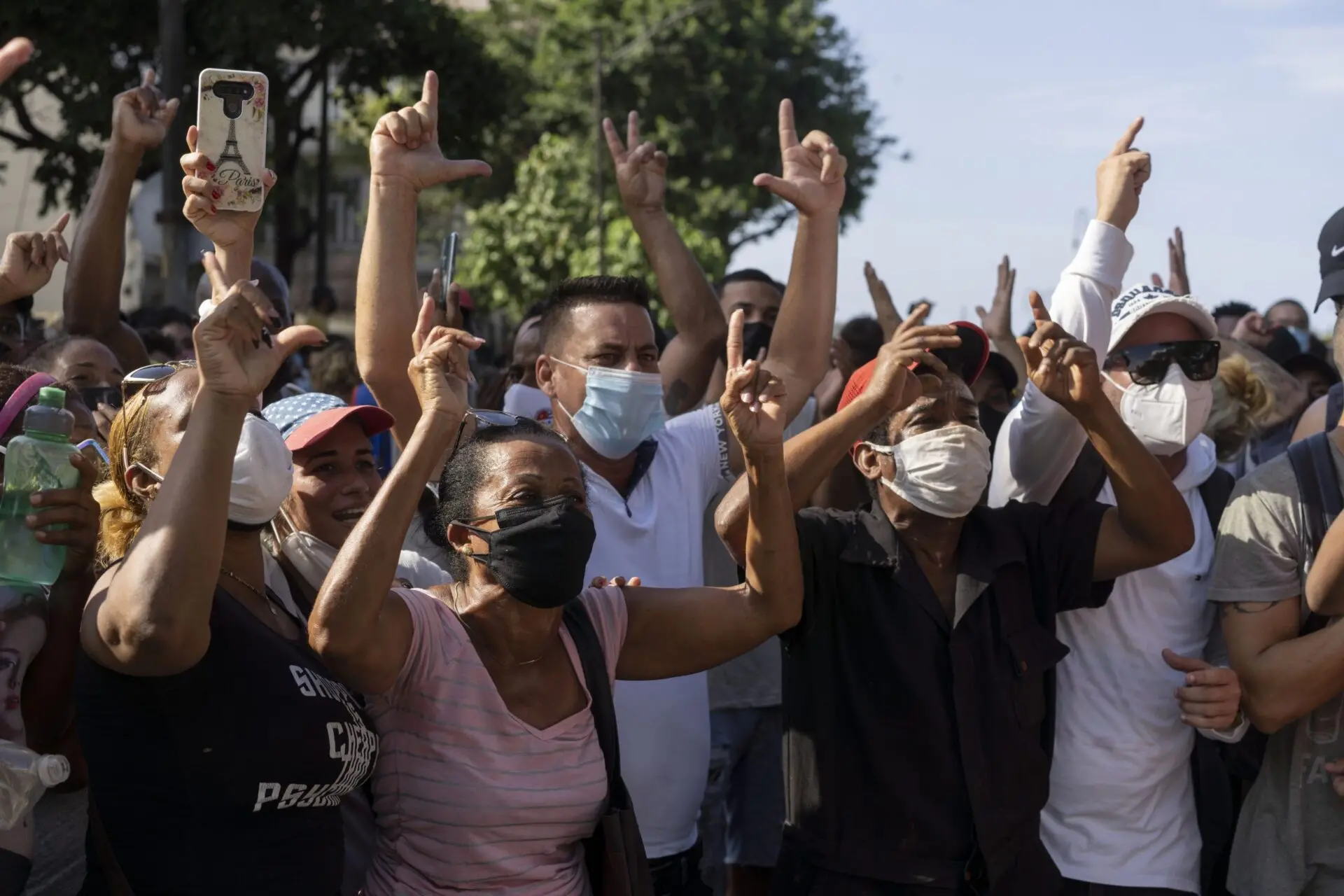 A group of people outdoors, many wearing face masks, raise their arms and hands in gestures, some making an L shape, appearing to protest or demonstrate during daytime. Trees and clear sky are visible in the background.