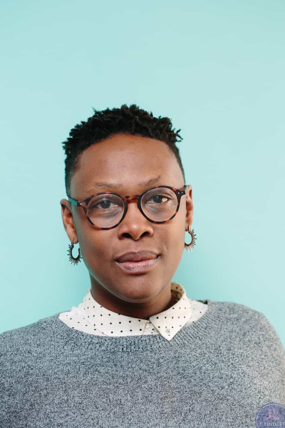 A person with short hair, round glasses, hoop earrings, and a grey sweater over a white collared shirt with black dots, stands against a light blue background.