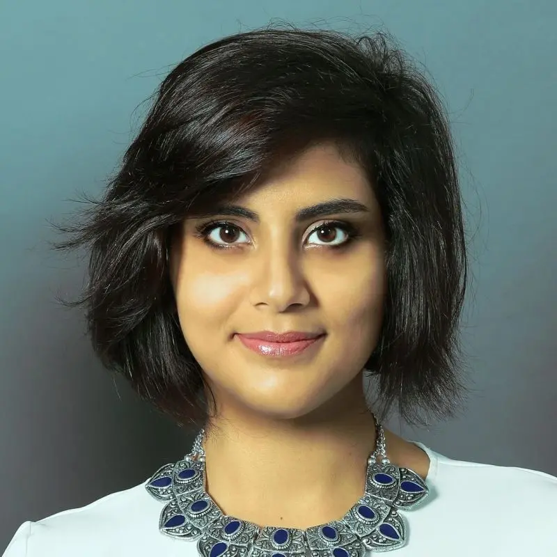 A woman with short, dark hair and brown eyes smiles softly. She wears a statement necklace with blue and silver details and a white top. The background is a solid, muted gray-blue color.