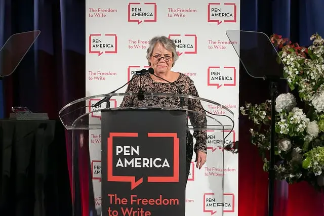 An older woman stands at a clear podium with a PEN America: The Freedom to Write sign, speaking at an event with a backdrop displaying the PEN America logo and a floral arrangement visible to the right.