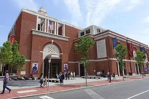 A large red-brick building with arched entrances, banners, and an American flag; people walk and sit outside. This is the Museum of the American Revolution in Philadelphia.