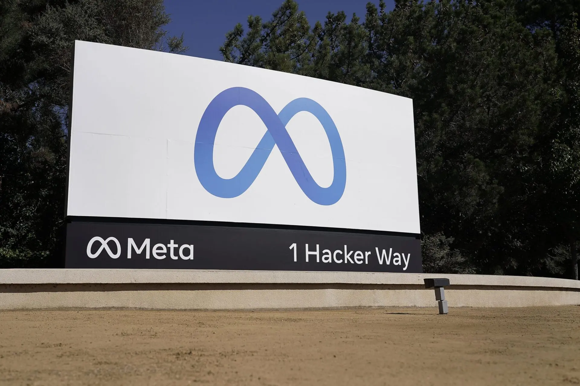 A large outdoor sign displays the Meta logo and text reading Meta and 1 Hacker Way, set against a backdrop of trees and clear sky.