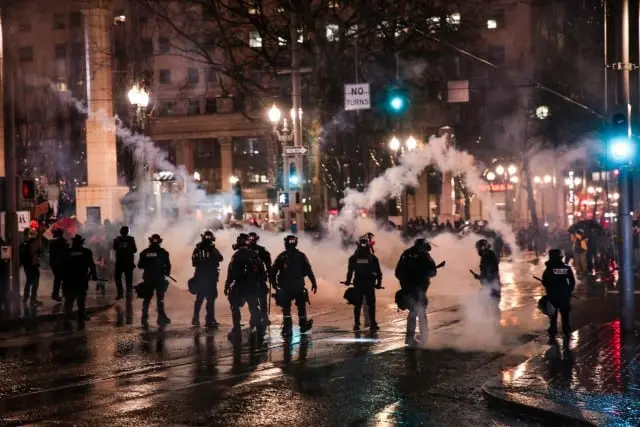 A line of police officers in riot gear face a crowd at night, with tear gas or smoke filling the air on a wet city street illuminated by streetlights.