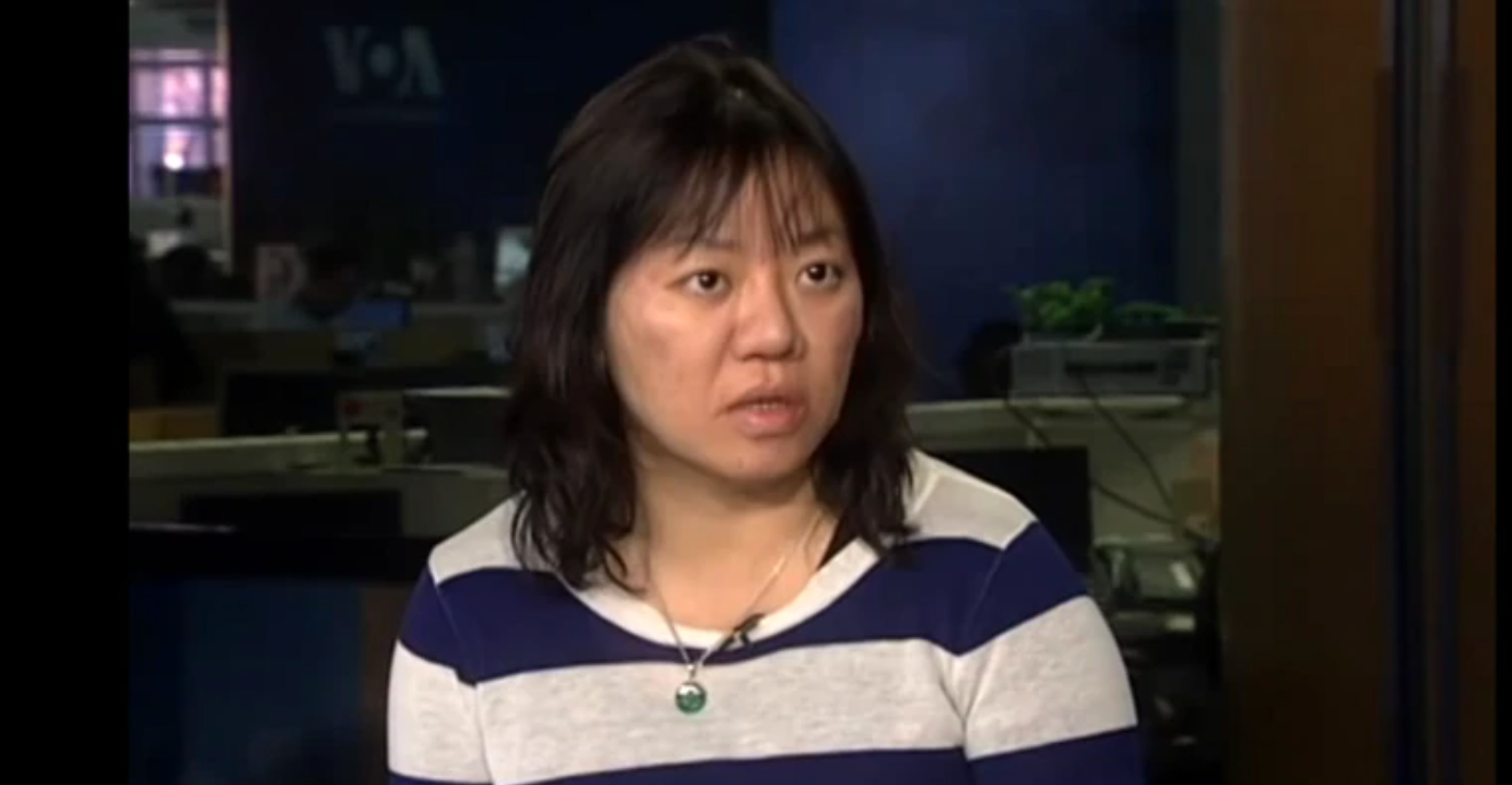 A woman with medium-length dark hair, wearing a navy and white striped shirt and a green pendant necklace, sits indoors with an office setting in the background.