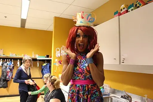 A person in a colorful dress and red wig, wearing a decorated paper crown, poses with a surprised expression in a classroom while two other people interact in the background.