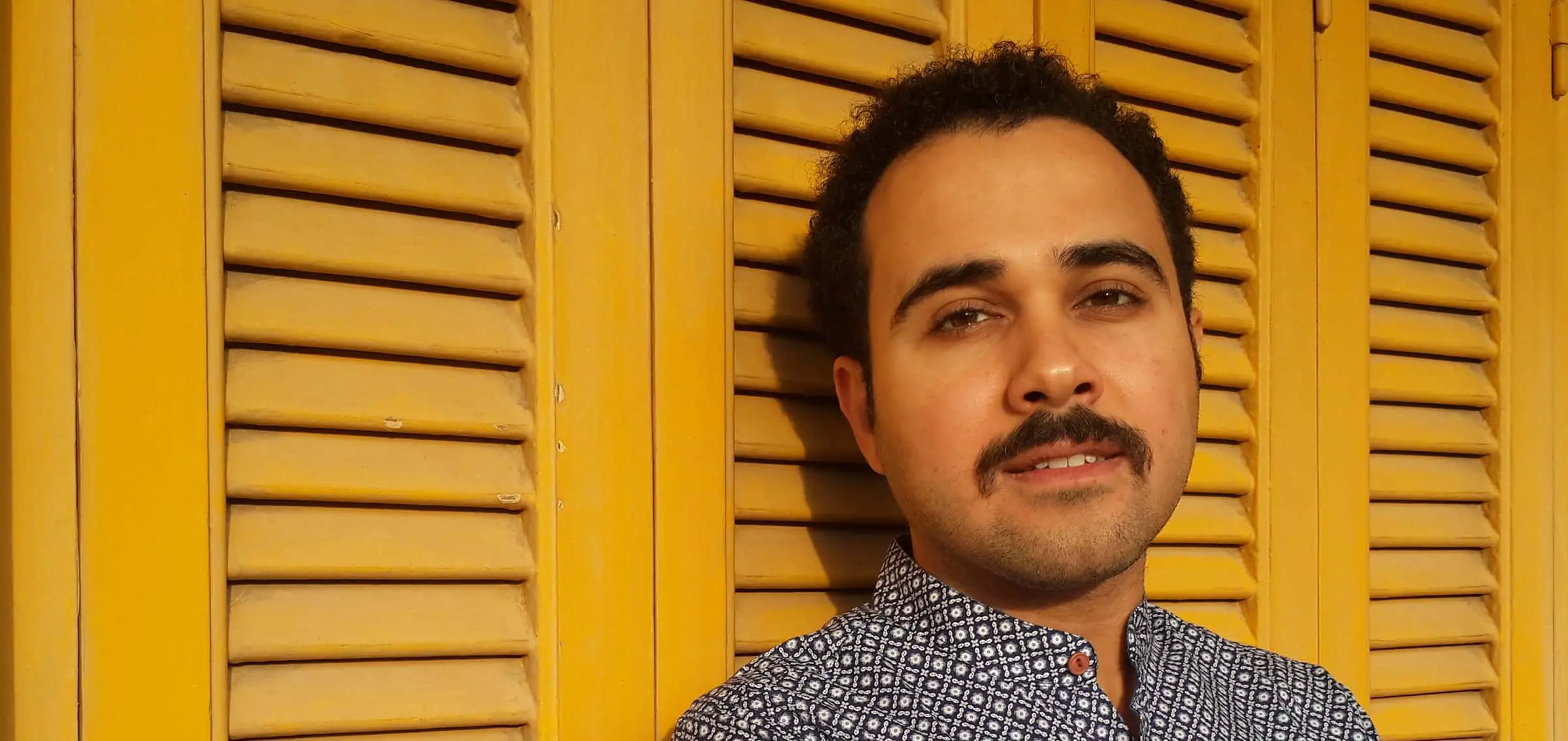 A man with short curly hair and a mustache stands in front of yellow wooden shutters, wearing a patterned button-up shirt and looking calmly at the camera.