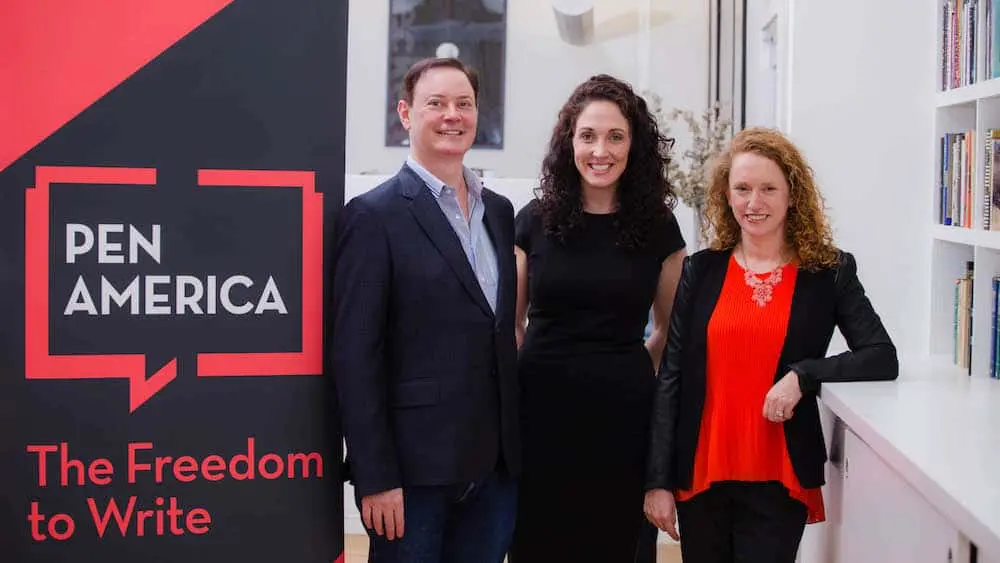 Three people stand smiling beside a large PEN America display sign that reads “The Freedom to Write” in a well-lit room with bookshelves in the background.