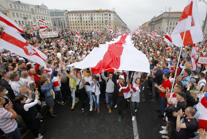 A large crowd of protesters fills a city street, many holding or wearing red and white flags. Several people in the center hold up a giant flag above their heads. The atmosphere is energetic and unified.