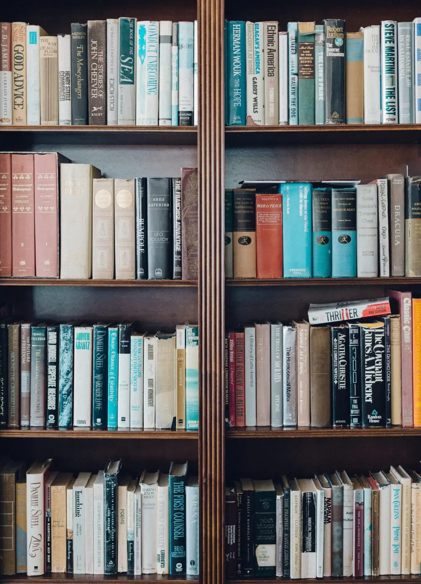 A wooden bookshelf filled with three rows of assorted books, including hardcovers and paperbacks in various colors, sizes, and genres. The books are tightly arranged side by side on both shelves.