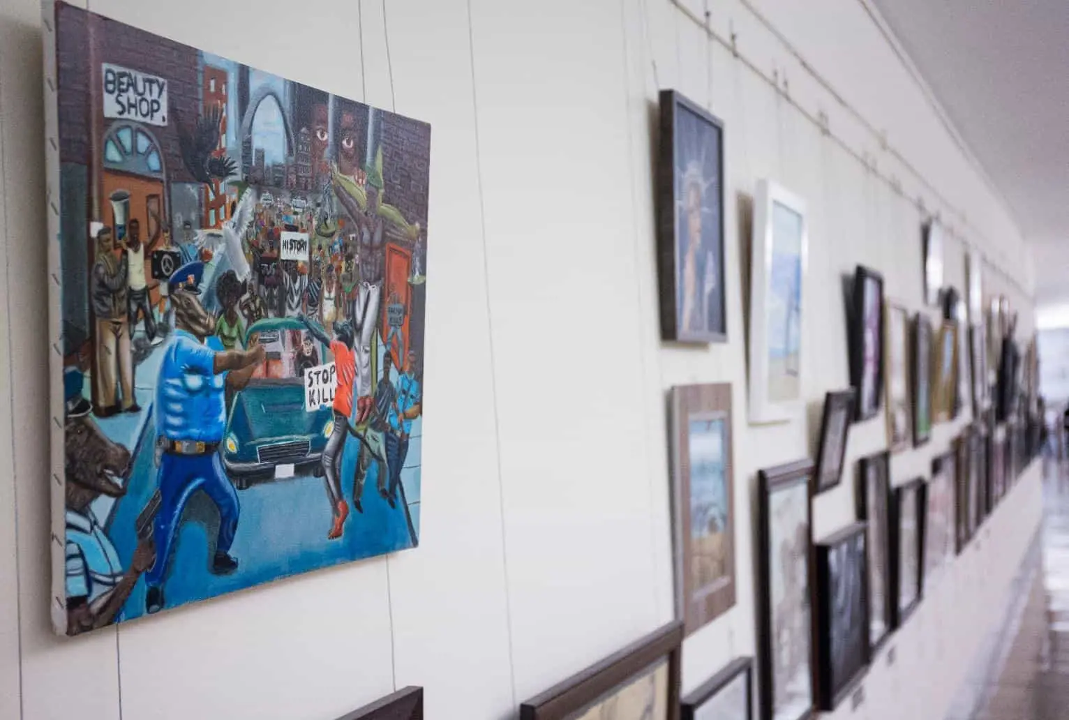 A hallway art gallery displays various framed artworks on a white wall, including a prominent painting depicting a police protest scene with people holding Stop Kill signs.