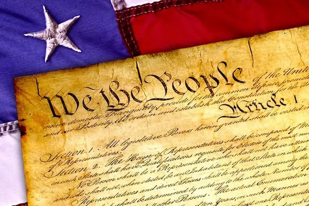 An old document with We the People written at the top, representing the U.S. Constitution, lies on an American flag with a visible white star.