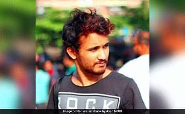 A young man with short, messy hair and a trimmed beard is standing outdoors. He is wearing a dark t-shirt with the word ROCK printed on it and is looking to his right. The background is blurred.
