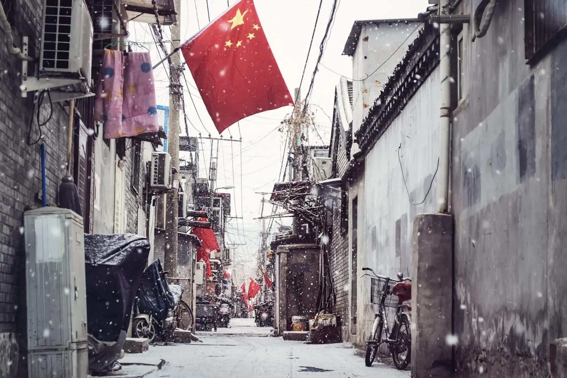 A narrow alleyway in China during snowfall, with bicycles and motorbikes parked along the sides and multiple Chinese flags hanging above. Old brick and concrete buildings line the street.