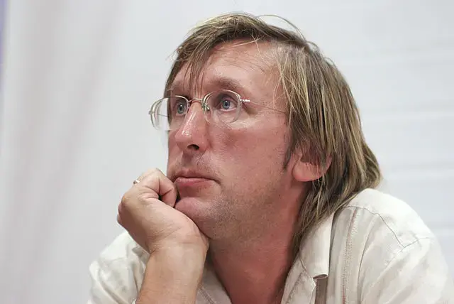A person with light brown hair and glasses rests their chin on their hand, looking thoughtfully into the distance against a plain, light background.