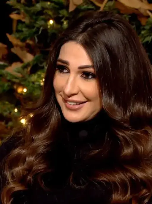 A woman with long, wavy brown hair and a black top smiles while sitting in front of a decorated, illuminated Christmas tree.