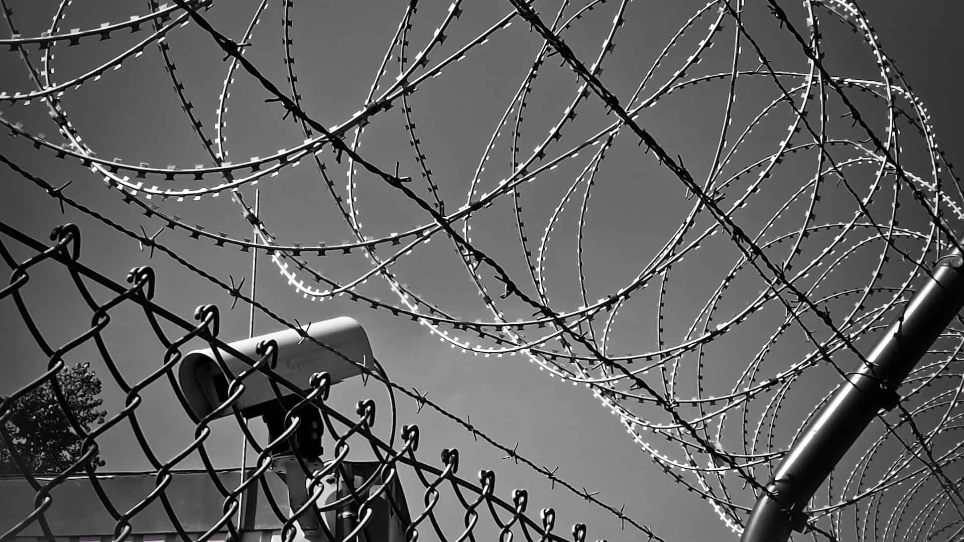 Barbed wire coils above a chain-link fence with a security camera mounted nearby, all set against a clear sky, conveying a sense of security and restricted access.