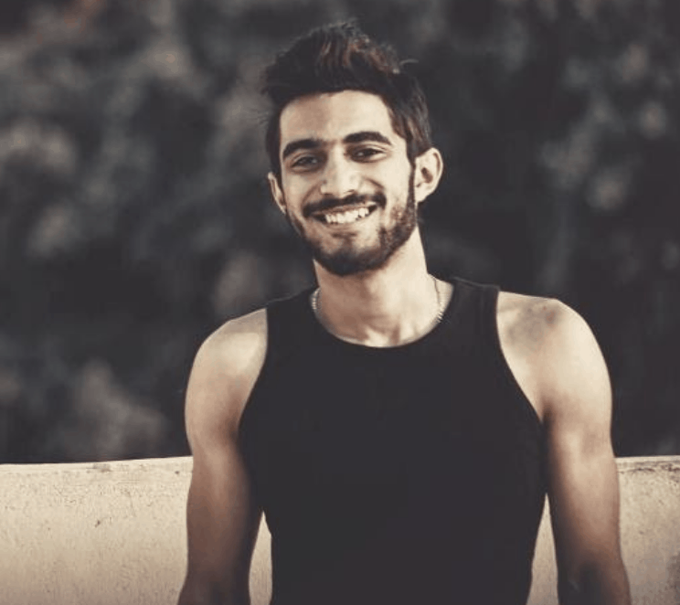 A young man with short dark hair and a beard, wearing a black sleeveless shirt, stands outdoors and smiles at the camera. The background is blurred with greenery.
