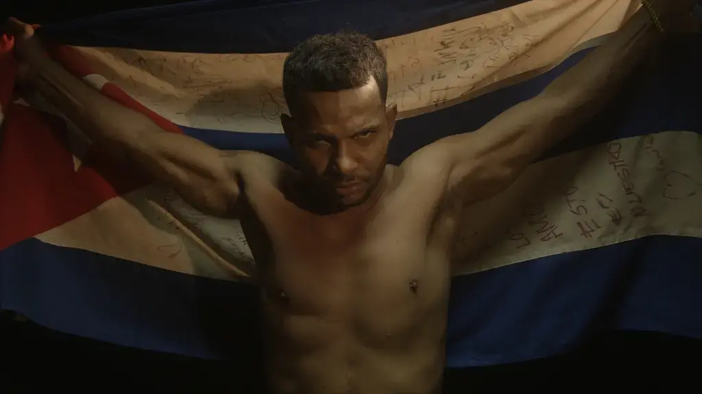 A shirtless man holds a large Cuban flag behind him with both hands, looking forward with a serious expression. The flag appears to have handwritten messages on it. The background is dark.