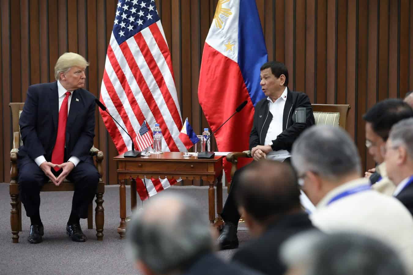 Former U.S. President Donald Trump and former Philippine President Rodrigo Duterte sit and talk in front of U.S. and Philippine flags during a formal meeting, with officials seated nearby.