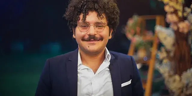 A smiling man with curly hair, glasses, and a mustache wears a navy blue suit and white shirt. He stands indoors with blurred floral decorations in the background.