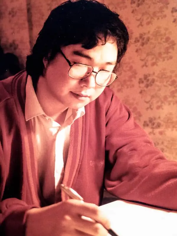 A person with glasses and dark hair, wearing a red jacket and white shirt, sits at a table under warm light with floral wallpaper, thoughtfully writing about conditional release.