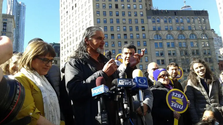 A man speaks at a podium surrounded by microphones during an outdoor rally, with people holding 32BJ SEIU signs and tall buildings in the background. Smiling supporters stand nearby.