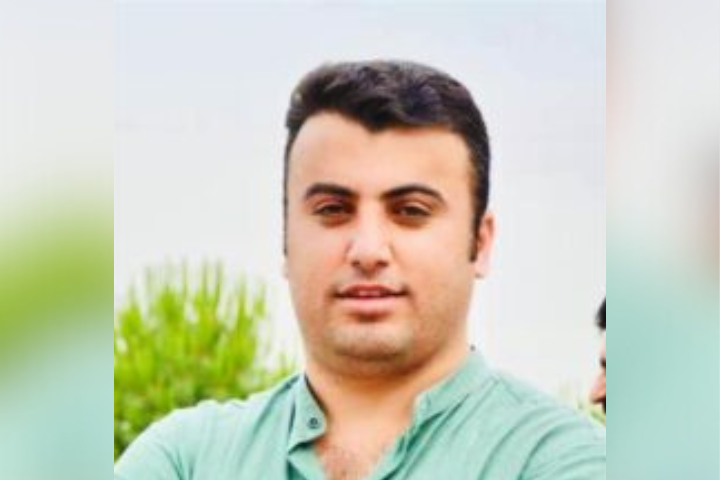 A man with short dark hair wearing a light green buttoned shirt stands outdoors with blurred greenery and a pale sky in the background. He is looking at the camera with a neutral expression.