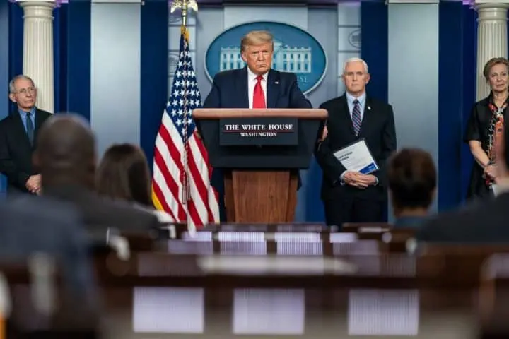 A man stands at a podium labeled The White House, flanked by officials, including a man holding papers, in a press briefing room reminiscent of Trump press conferences, with US flags and a seated audience in the foreground.