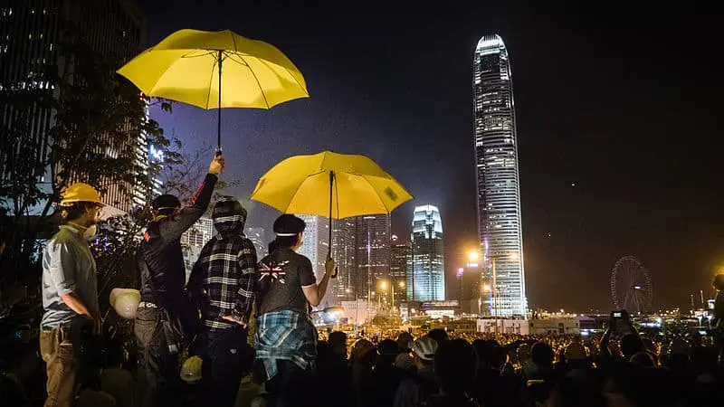 A group of people hold bright yellow umbrellas at night, facing a city skyline with tall, illuminated skyscrapers. The scene is crowded, and city lights glow in the background.