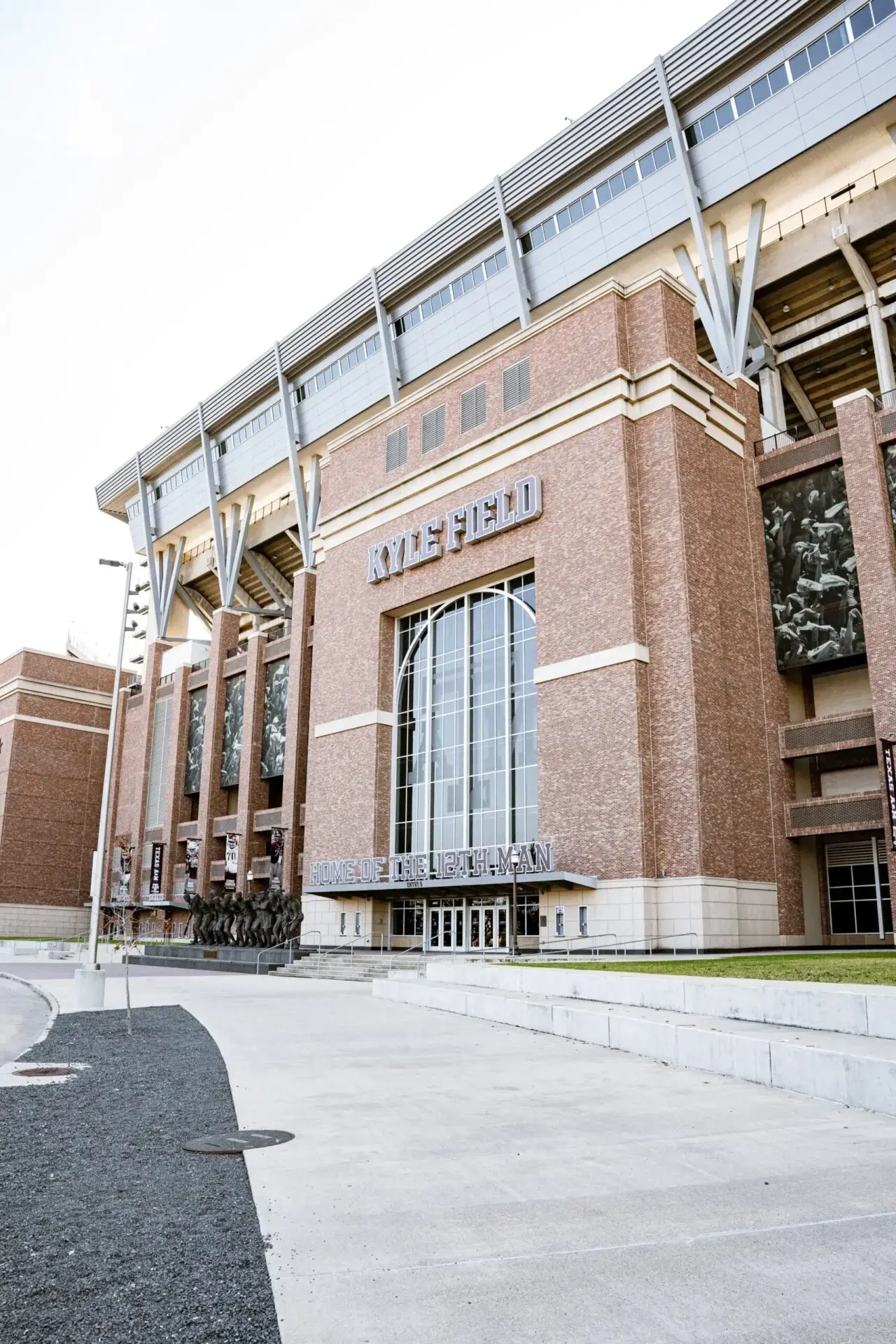 Exterior view of Kyle Field, a large brick football stadium at Texas A&M University, with visible statues and signage near the entrance on a bright day.
