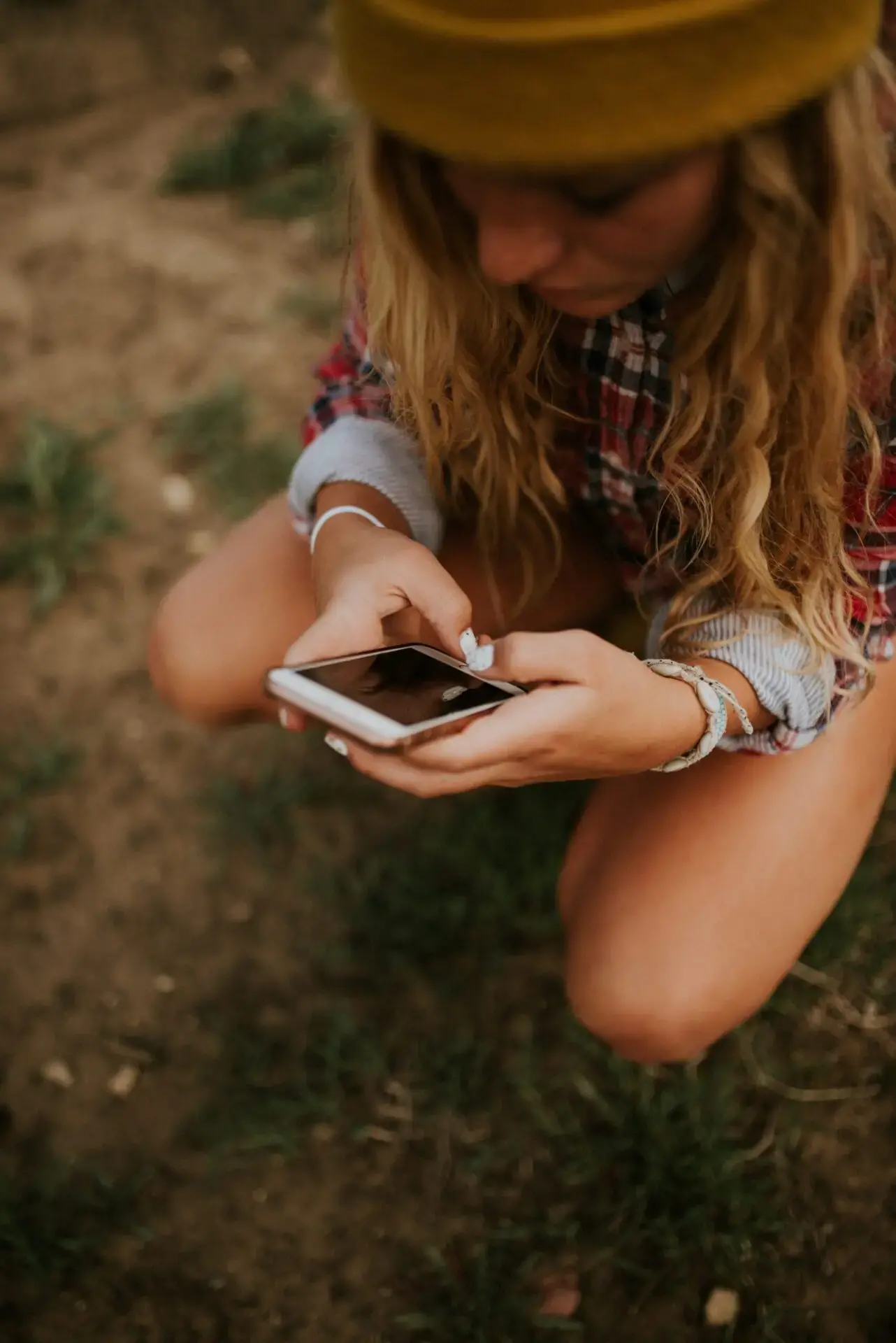 A person with long blonde hair, wearing a plaid shirt and sitting outdoors on the ground, is looking down at and using a smartphone. The background is earthy with patches of grass.