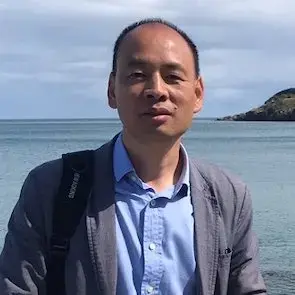 A man wearing a blue shirt and gray jacket stands in front of a calm body of water with a backpack over one shoulder. The sky is cloudy and a small hill is visible in the background.