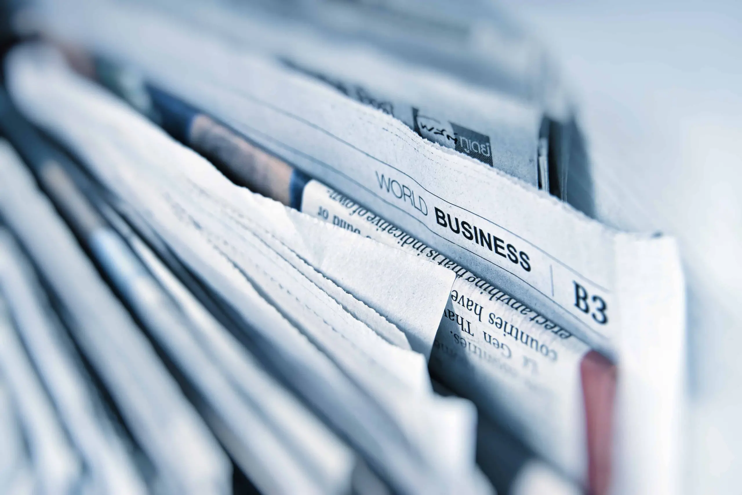A close-up of several folded newspapers stacked together, with a visible section titled World Business and the page number B3 in focus. The image is in cool blue tones.