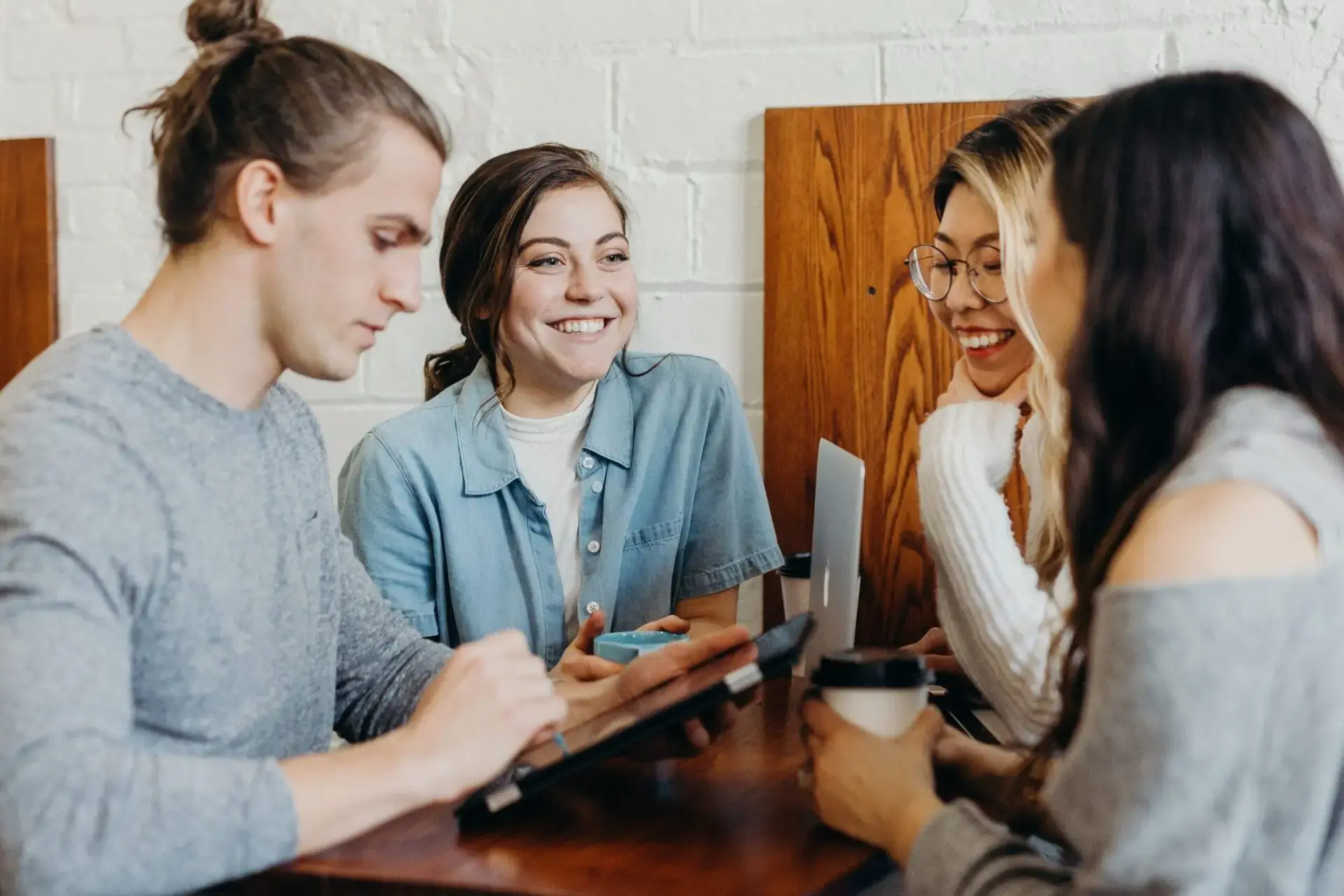 Four young adults sit at a wooden table, talking and smiling. One uses a tablet, another holds a coffee cup. They appear to be having a friendly conversation in a casual indoor setting.