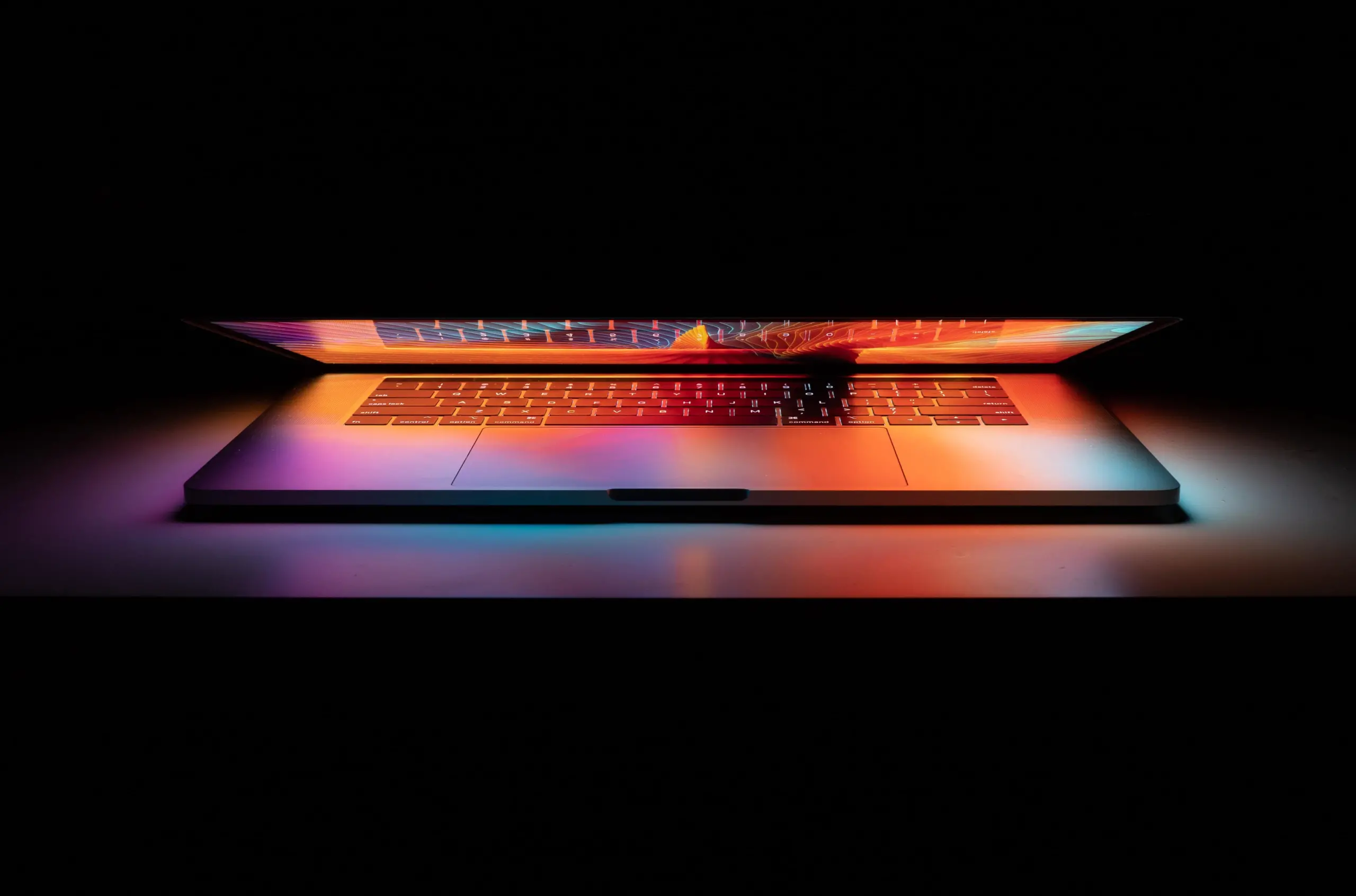 A partially open laptop glows with vibrant, colorful light from its screen and keyboard, creating a dramatic effect against a dark background.