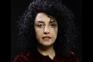 A person with curly black hair and light skin, wearing a dark red velvet top, is facing the camera against a plain black background.