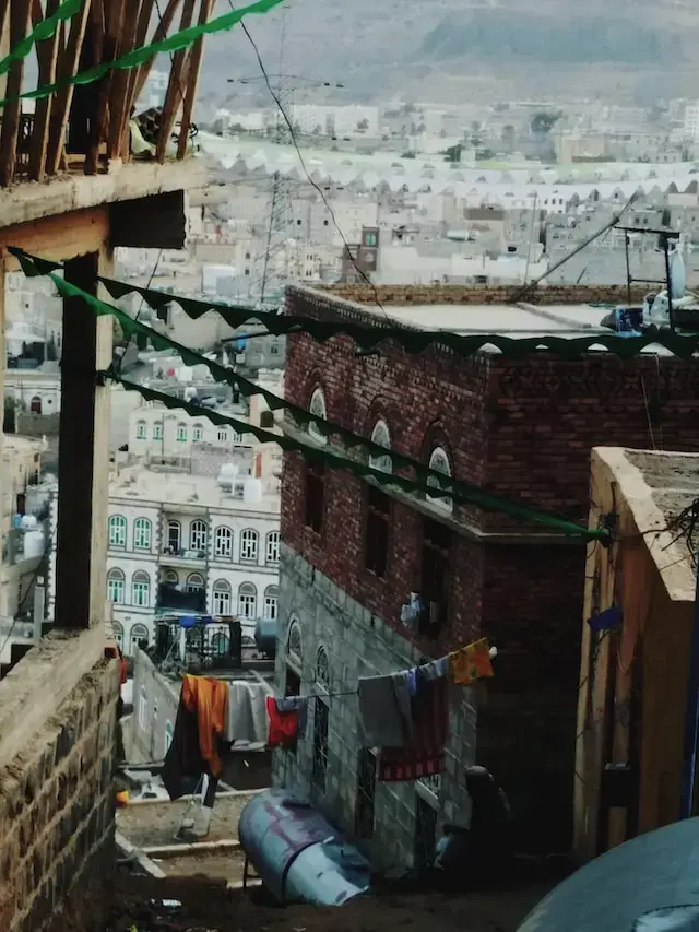 A narrow alley between brick buildings, with green banners strung overhead and laundry hanging on lines. Cityscape and mountains are visible in the hazy background.