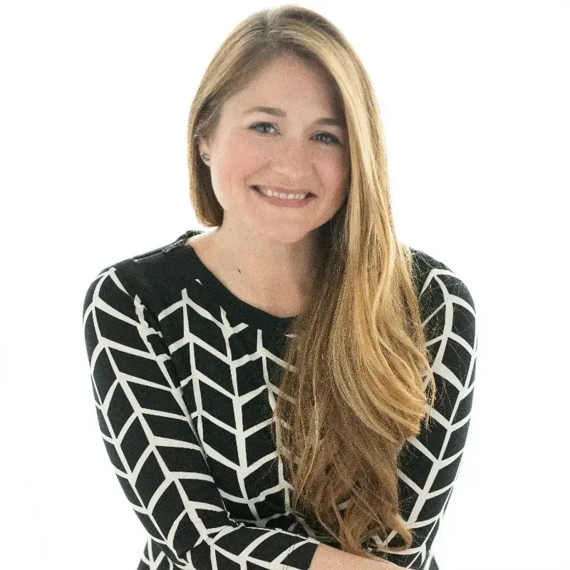 A woman with long, light brown hair wearing a black and white patterned top smiles softly in front of a white background.