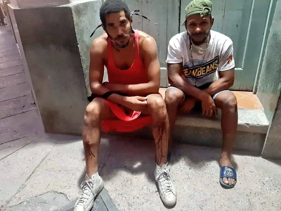 Two men sit on a concrete bench. The man on the left wears a red sleeveless outfit and sneakers, with marks on his legs. The man on the right wears a cap, white shirt, shorts, and a single blue sandal. Both look toward the camera.