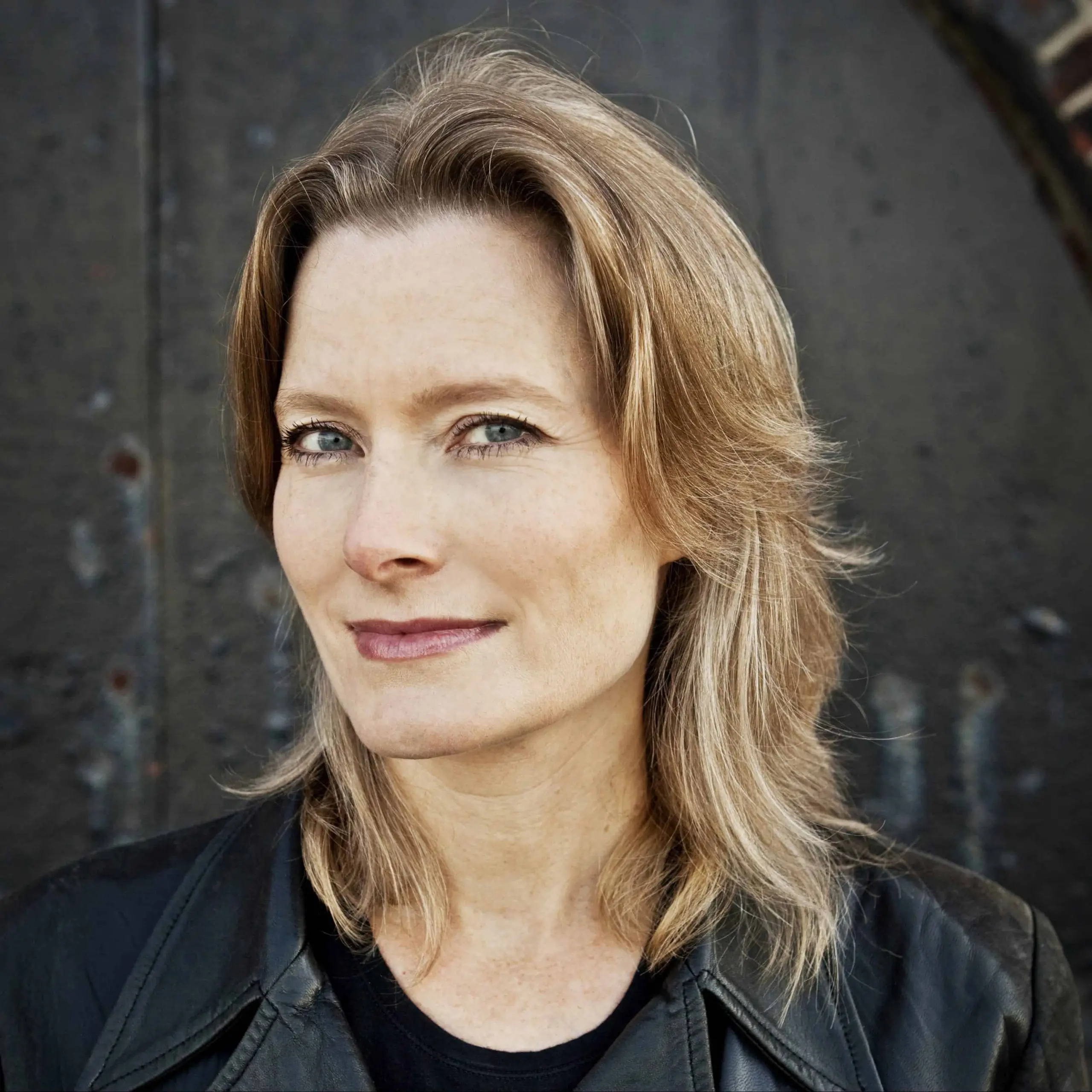 A woman with shoulder-length light brown hair and blue eyes smiles slightly while wearing a black leather jacket, standing in front of a dark, textured background.
