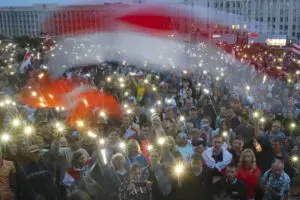 A large crowd of people gathered outdoors at dusk, holding up lit phones and waving a red and white flag, creating a sea of lights and color in front of a large building.