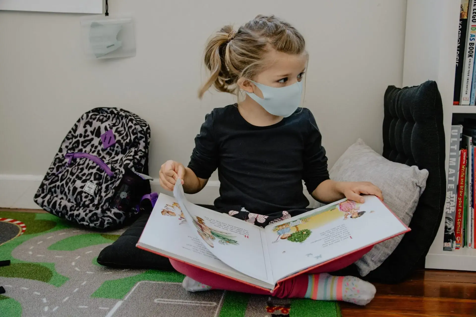 A young girl wearing a face mask sits on the floor, reading a picture book. She is surrounded by cushions, a backpack, and a shelf with books, with part of a play mat visible under her.