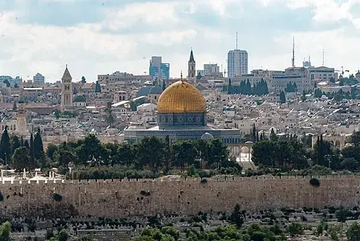 A panoramic view of Jerusalem with the golden Dome of the Rock prominently in the center, surrounded by historic buildings, trees, and cityscape under a partly cloudy sky.